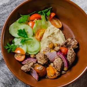 Shawarma spiced chicken with roasted tomatoes and onions, plus a dollop of hummus and some cucumber salad in a brown bowl on a linen napkin.
