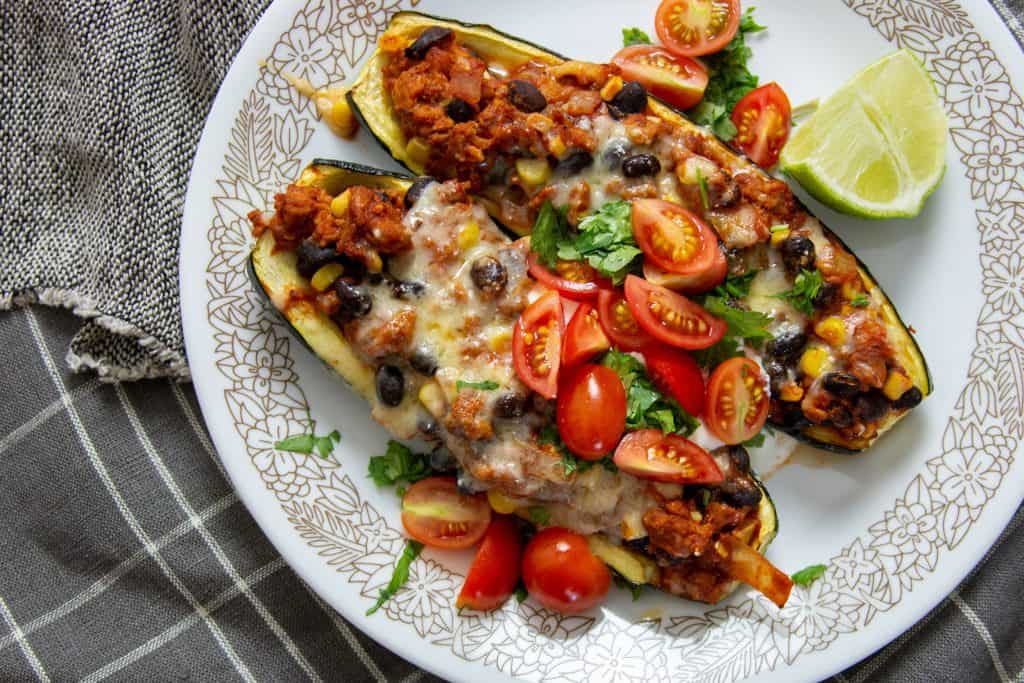 Close up of two turkey taco zucchini boats on a plate with fresh herbs, tomatoes, and a wedge of lime.