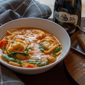 A bowl of tomato tortellini soup on a table with a chunk of parmesan cheese, a bottle of balsamic vinegar, and a checkered napkin in the background.