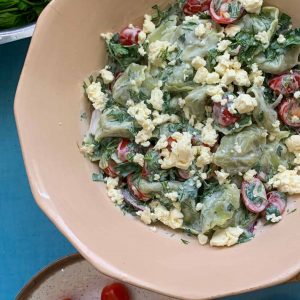 A large bowl of tortellini salad with a bowl of cherry tomatoes on a blue table.