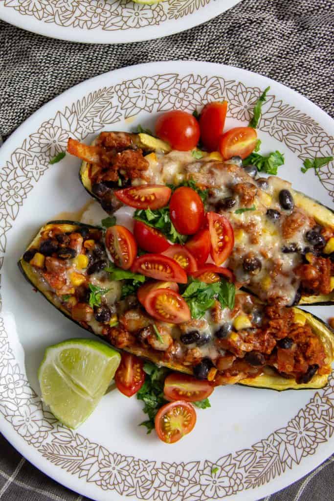 Two turkey taco zucchini boats on a plate with fresh herbs, tomatoes, and a wedge of lime.