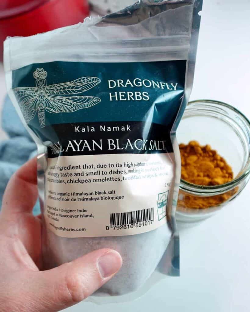 A woman holding a bag of black salt with a jar of turmeric in the background.