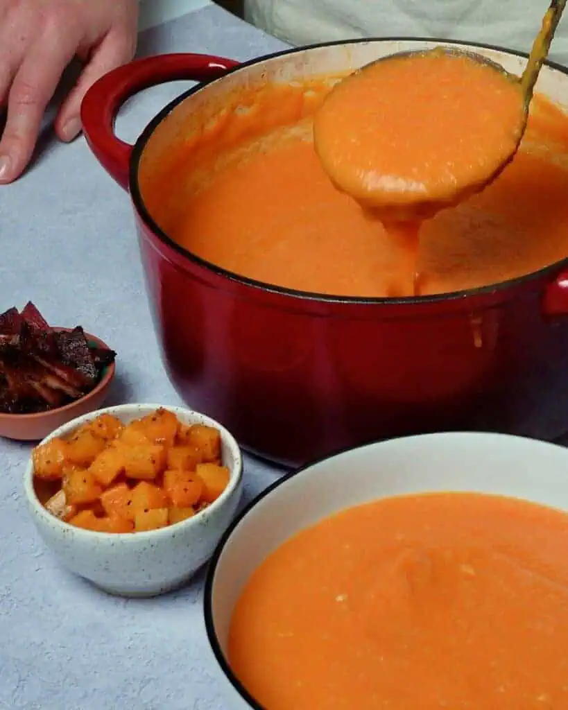 A woman ladling carrot butternut soup into a bowl. Cubed butternut squash and bacon in small bowls.