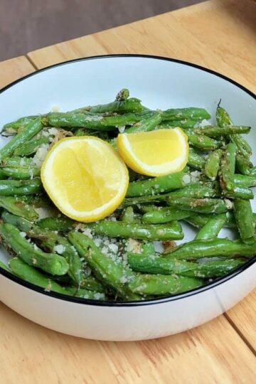 garlic parmesan green beans in a white bowl with lemon wedges on top.