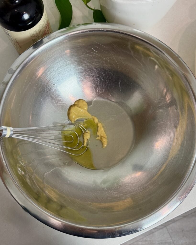 Whisking dijon mustard, lemon juice, and olive oil in a stainless steel bowl.