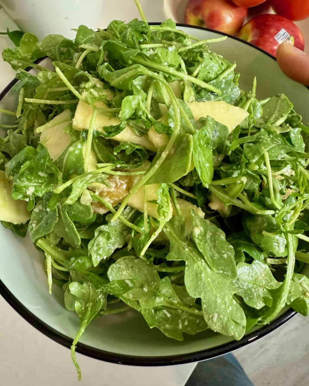 A woman holding a bowl of arugula salad with apples in the background.