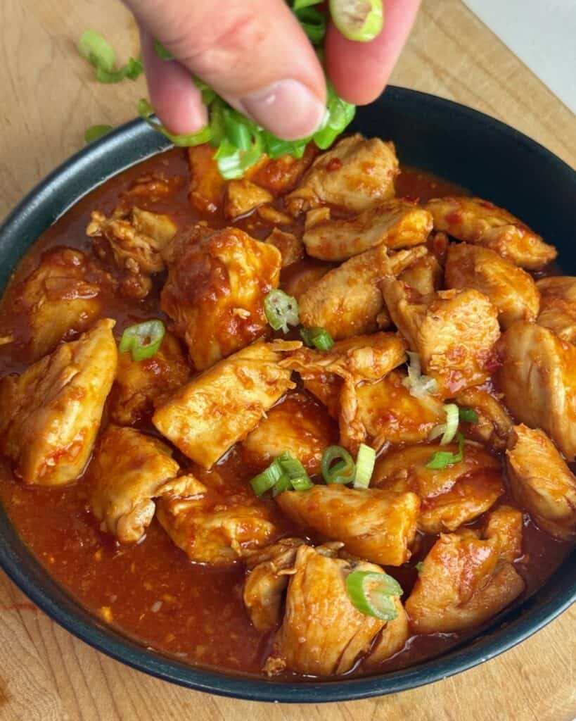 A woman sprinkles gochujang chicken with sliced green onions.