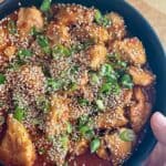 A woman holds a plate of gochujang chicken with sesame seeds and green onions.