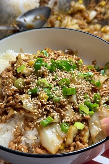 A woman holds pork and cabbage stir fry over rice in a white bowl with black trim.
