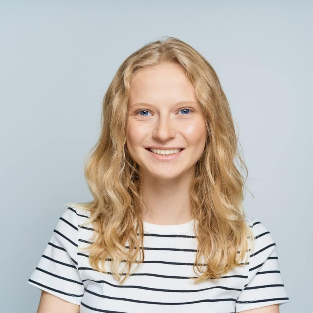 Smilende kvinde med blonde hår og blå øjne mod lyseblå baggrund.