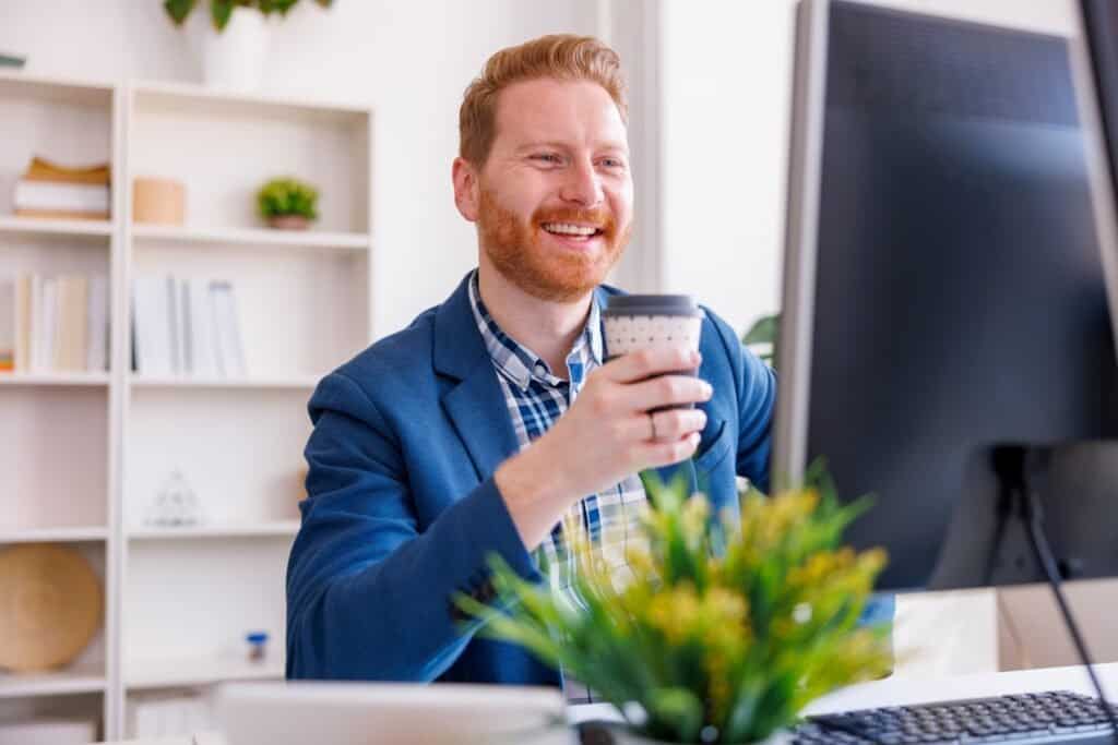 Berømt mand smiler med kaffe foran computer på kontor.