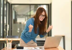 Glad kvinde fejrer succes foran sin laptop på arbejdspladsen.