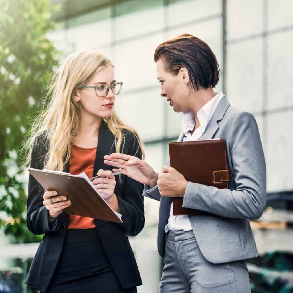 To kvinder i professionel samtale udenfor, med mapper i hænderne.