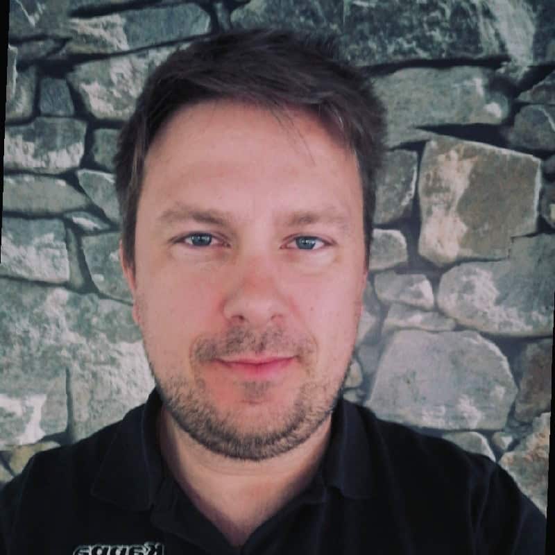 Mand med kort hår foran en stenmur, smilende, i sort skjorte.