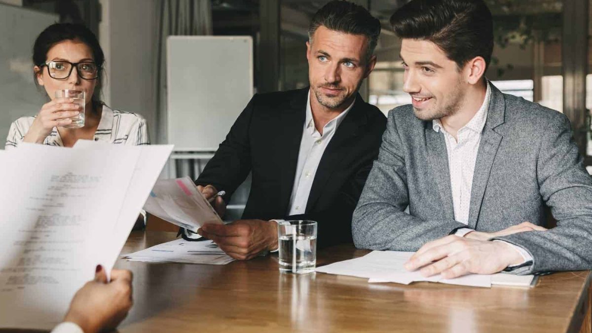 Kvindelig medarbejder drikker vand under møde med to mænd i forretningsmiljø.