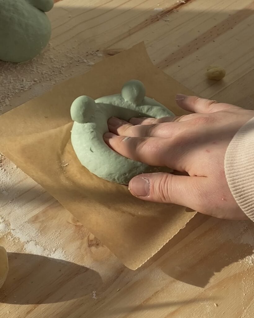 Uncooked frog-shaped pizza dough, formed into the shape of a frog with raised edges for the body and small dough balls for the eyes, ready to be topped with cheese and other ingredients before baking.