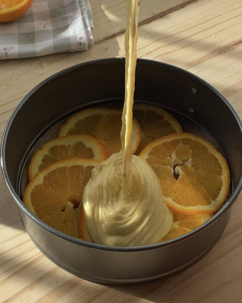 Thinly sliced oranges arranged on parchment paper in a baking pan with orange cake batter poured on top, creating a perfect base for an upside-down orange cake.