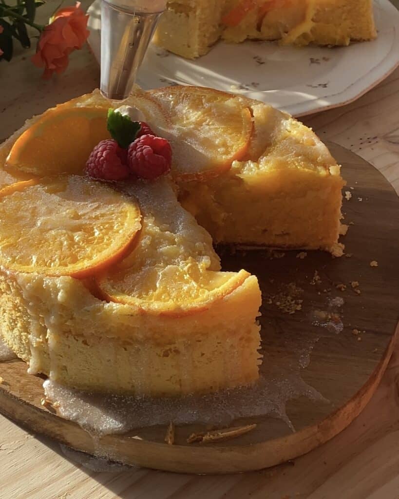 Close-up of a moist upside-down orange cake being decorated with piped swirls of heavy cream, accented with fresh raspberries and mint leaves, showcasing a vibrant and elegant presentation with a glossy orange glaze for a citrusy finish