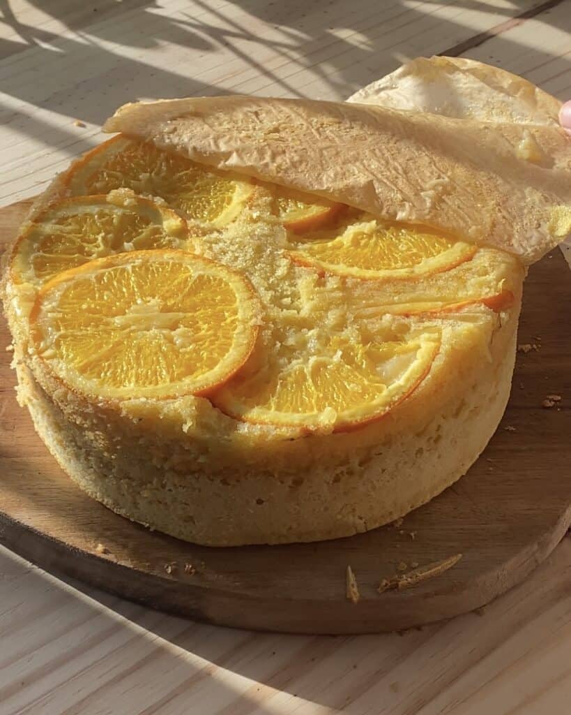 Gently peeling off parchment paper from the freshly baked upside-down orange cake, revealing perfectly arranged caramelized orange slices on top of the moist citrus cake.