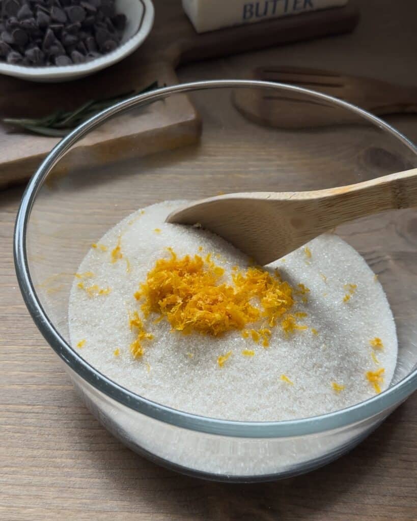Rubbing fresh citrus zest with sugar, releasing aromatic oils to infuse the mixture with vibrant flavor for the chocolate chip muffins