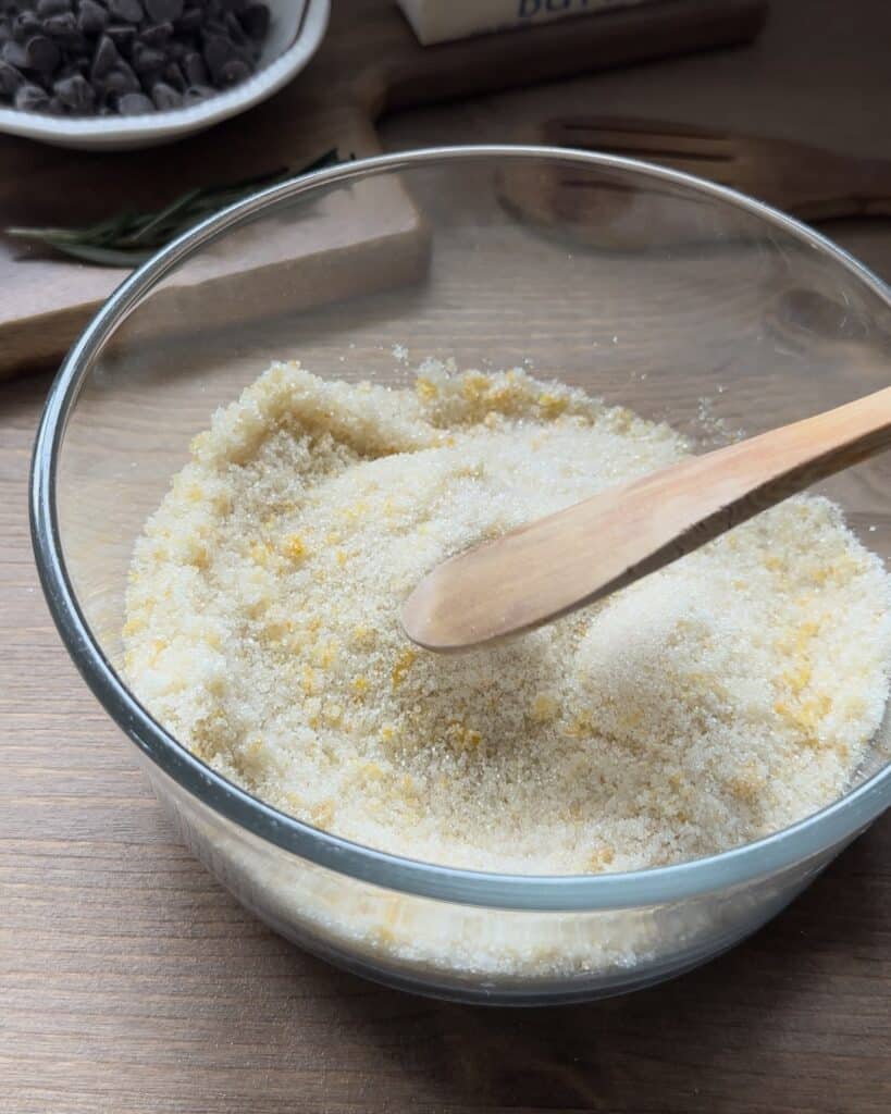 Rubbing fresh citrus zest with sugar, releasing aromatic oils to infuse the mixture with vibrant flavor for the chocolate chip muffins