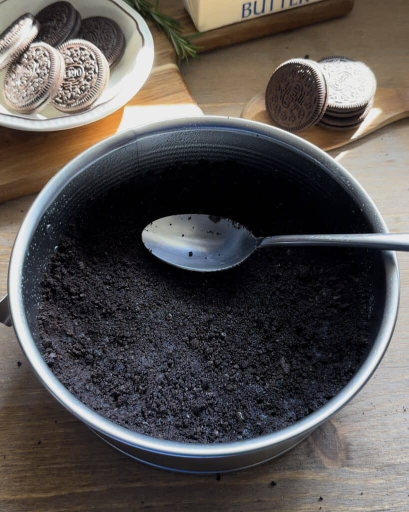 Adding Oreo crumbs mixed with melted butter into a greased pan, pressing them down to form the crust for the no-bake Oreo cheesecake
