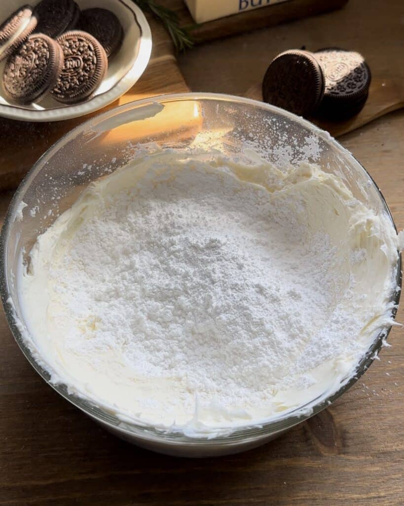 Adding sugar to softened cream cheese, mixing them together to create a smooth and sweet filling for the no-bake Oreo cheesecake