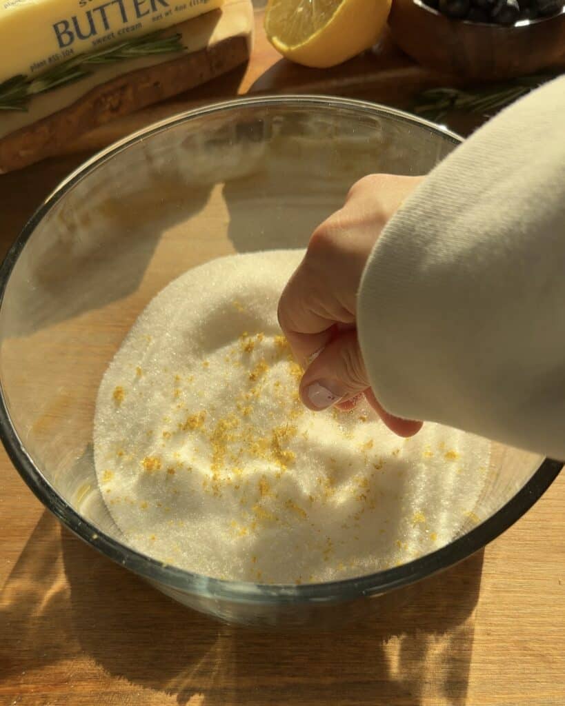 Infusing sugar with fresh lemon zest by rubbing them together to create a zesty base for bakery-style blueberry lemon muffins