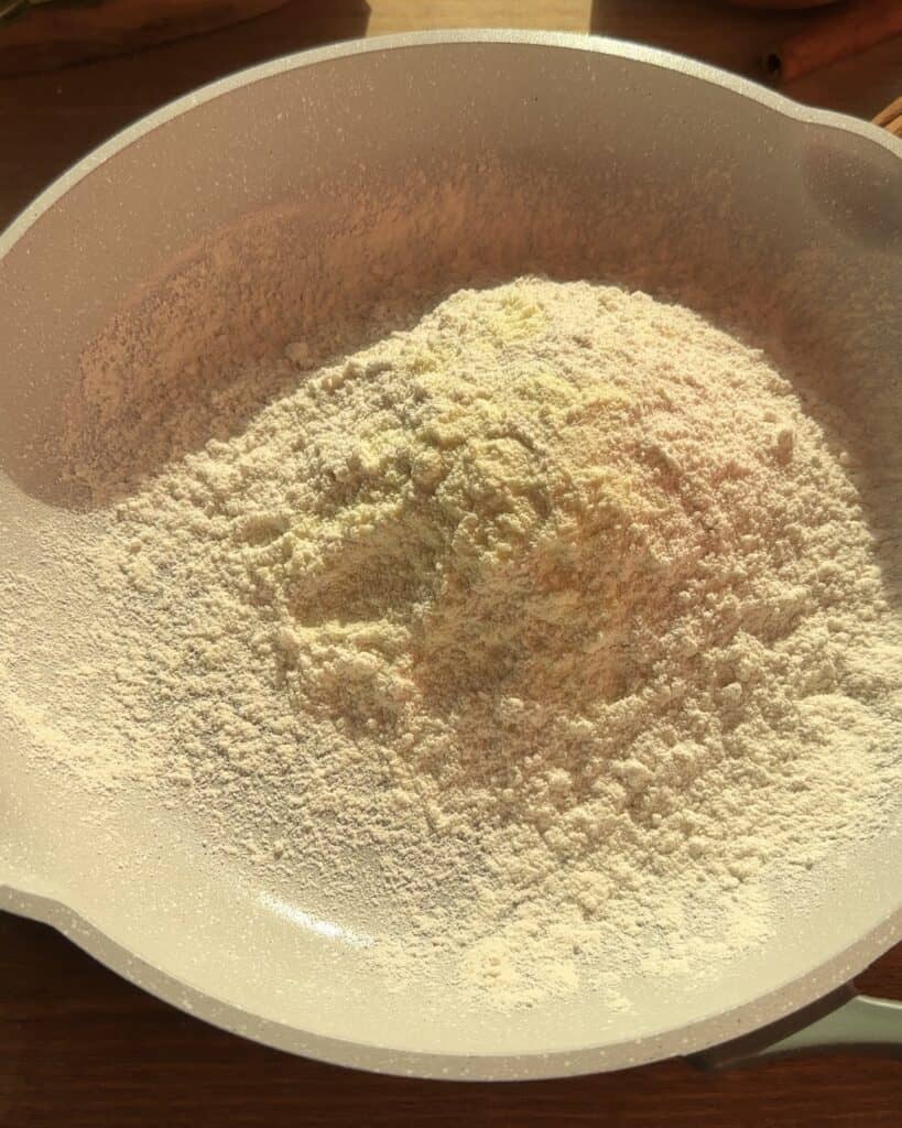 Toasting flour and dry milk powder in a saucepan to enhance the flavor for the Christmas cookie dough cheesecake batter.