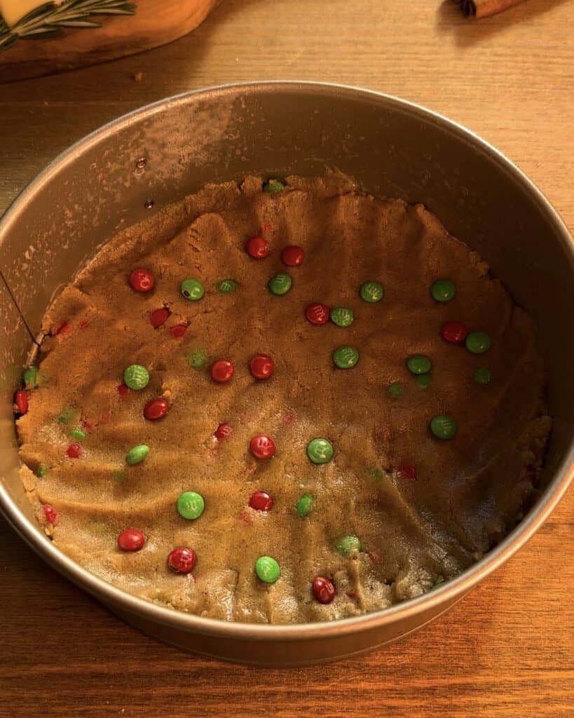 Spreading 3/4 of the cookie dough evenly in a greased baking pan to create the crust for a Christmas cookie dough cheesecake, packed with festive red and green M&M’s.