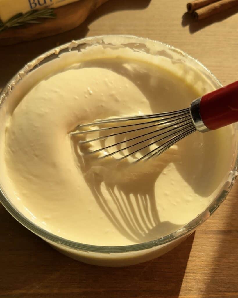 Mixing cream cheese, granulated sugar, heavy whipping cream, and eggs with a whisk in a bowl to create a smooth and creamy batter for the Christmas cookie dough cheesecake.