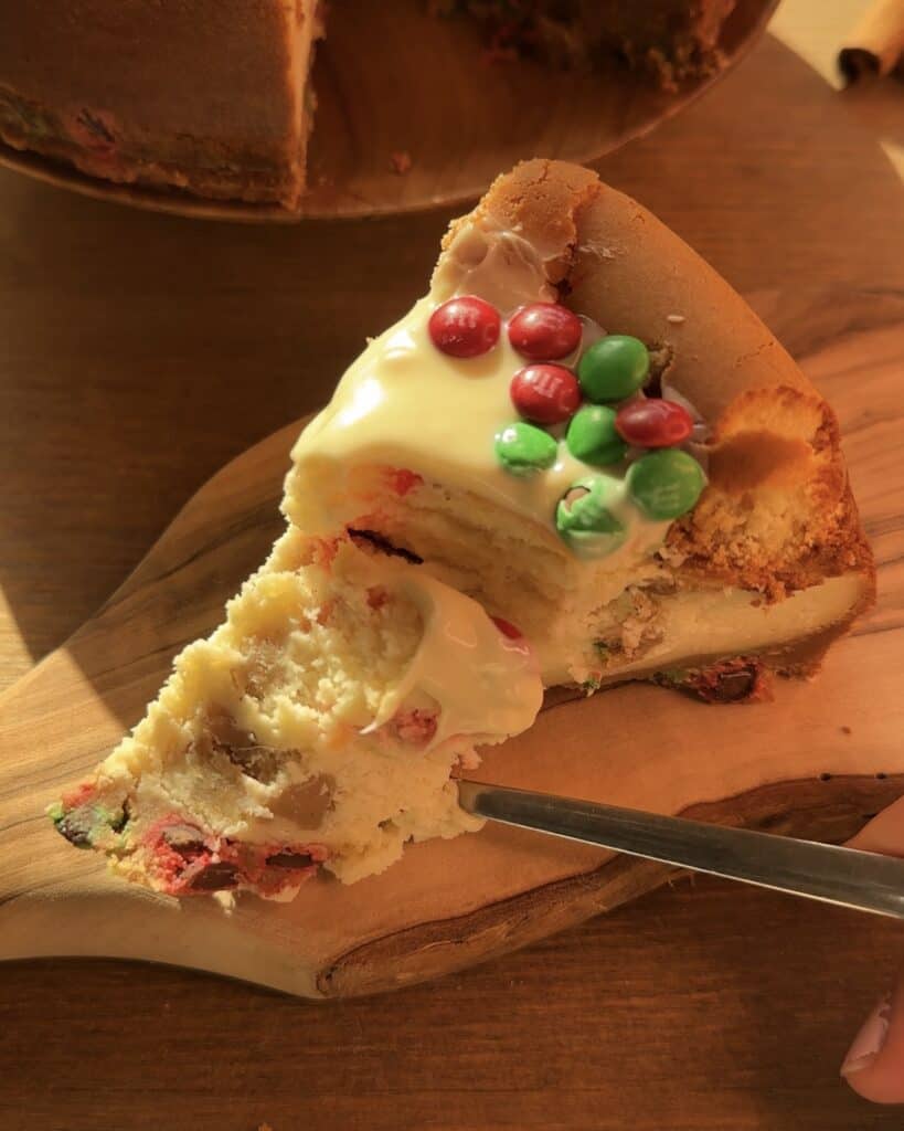 A slice of Christmas cookie dough cheesecake opened with a spoon, revealing its creamy interior. It features a cookie dough crust, rich cheesecake filled with red and green M&M’s, and a topping of melted white chocolate and more M&M’s, creating the perfect festive dessert.