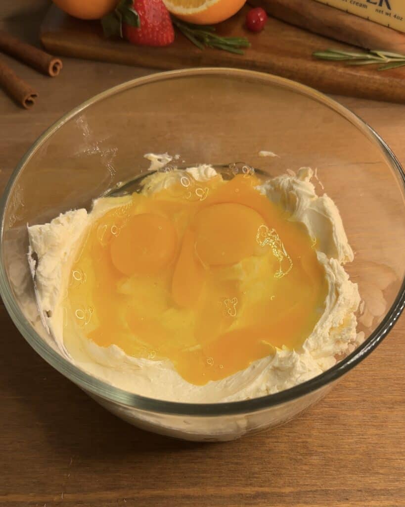Adding eggs and sugar to the creamy, whipped cream cheese in a bowl, preparing the filling for Churro cheesecake bars