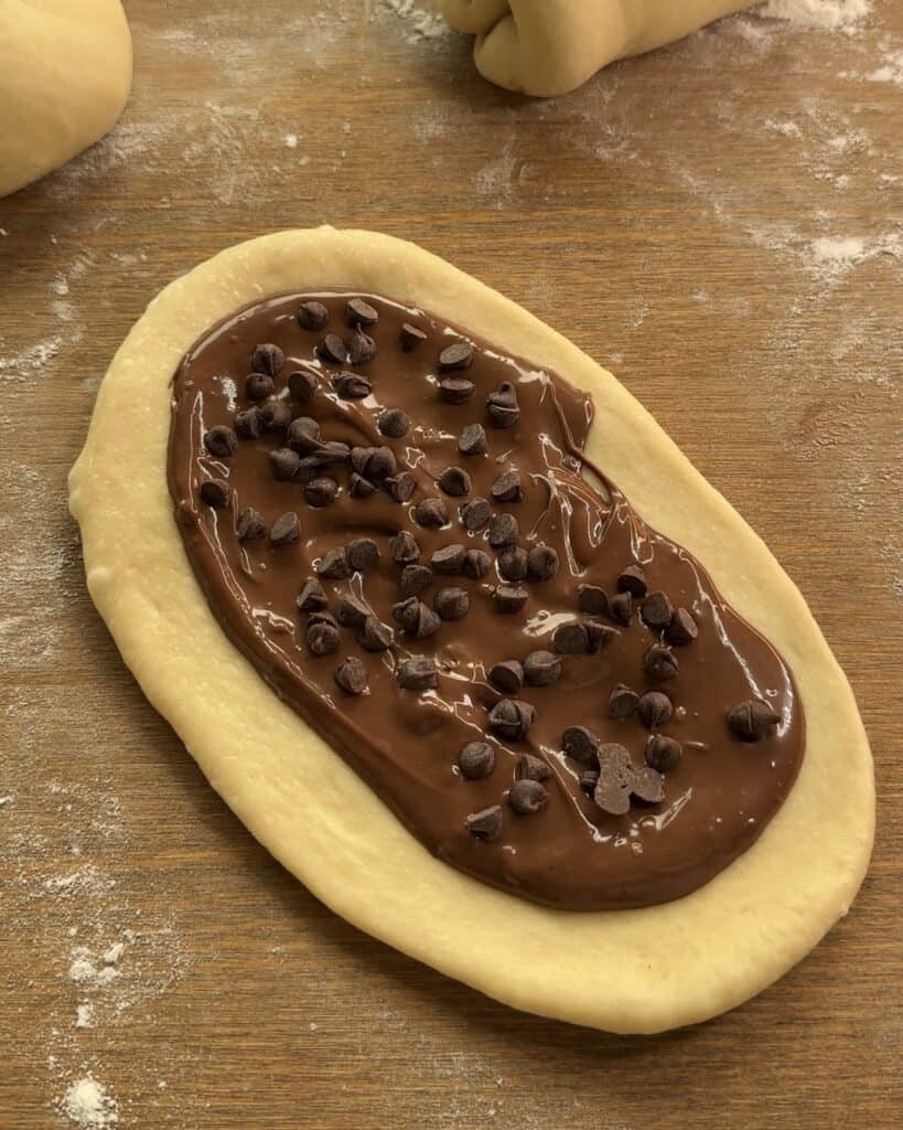 Flattening each dough ball into an oval shape and spreading Nutella in the center using a rolling pin to prepare Nutella-stuffed brioche