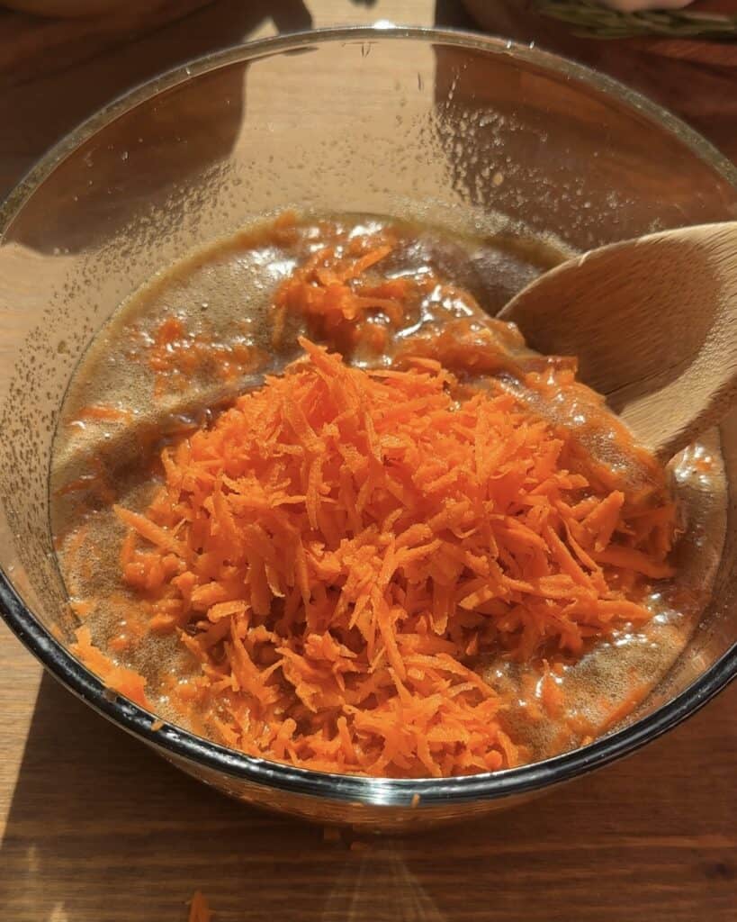 Freshly grated carrots resting on top of the mixed wet ingredients, ready to be folded into the carrot cake batter for a moist and flavorful texture.