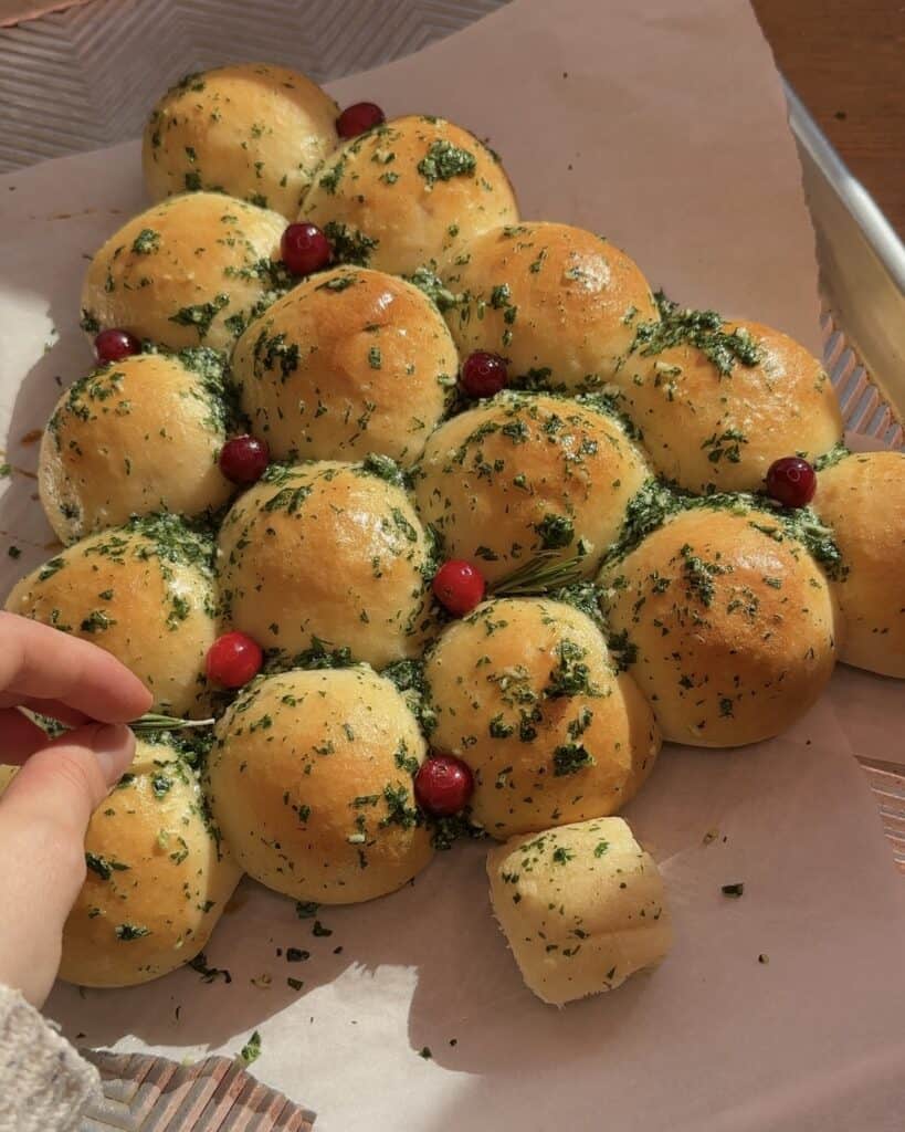 Adding cranberries as ornaments, rosemary as tree branches, and Parmesan cheese as snow to the pull-apart Christmas tree garlic bread, creating a festive and flavorful decoration.