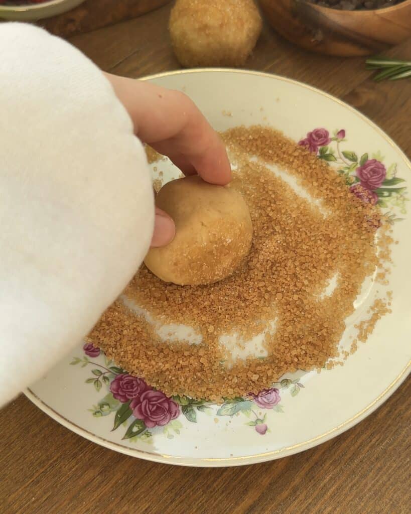 A close-up of a hand rolling a dough ball in coarse brown sugar on a decorative floral plate, preparing for baking.