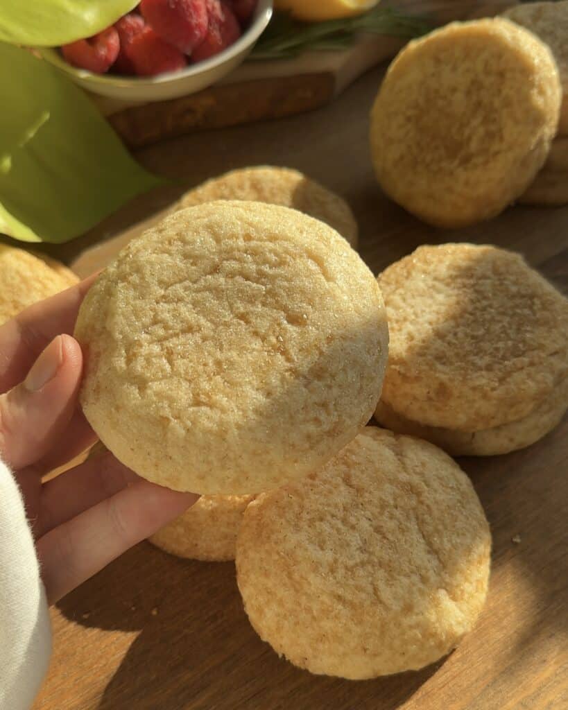 Golden, freshly baked snickerdoodle cookies with a sugar-coated crust, displayed under natural sunlight with a bowl of fresh raspberries in the background.