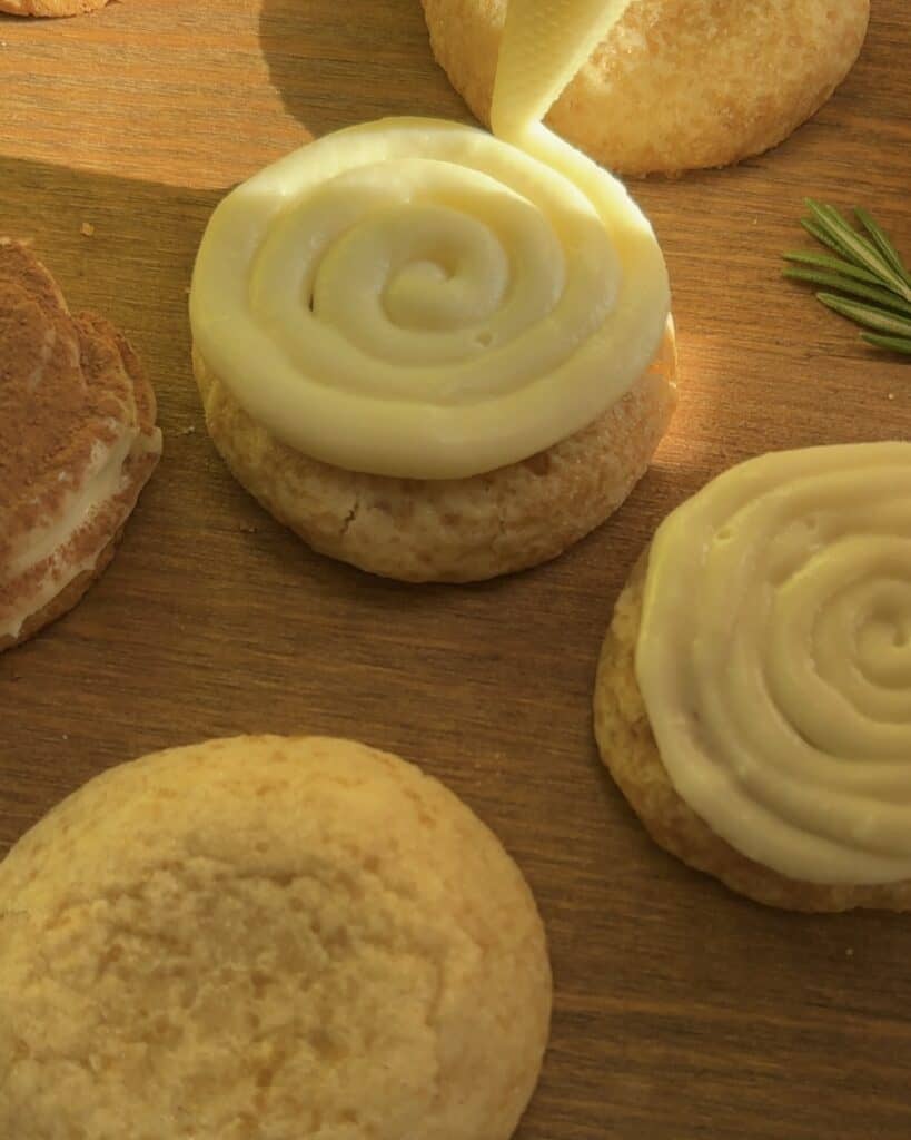 A piping bag swirling cream onto a cookie, surrounded by plain and partially assembled cookies on a wooden surface