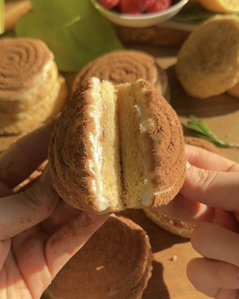 Hands holding a tiramisu cookie split in half, revealing a creamy filling sandwiched between two soft cookie layers with more cookies and greenery on the background