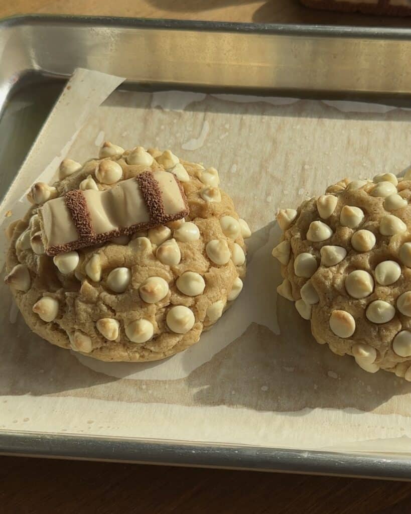 Two freshly baked Kinder Bueno cookies on a baking pan, one with a piece of Kinder Bueno in the center, and the other without, showing the difference in appearance before serving.