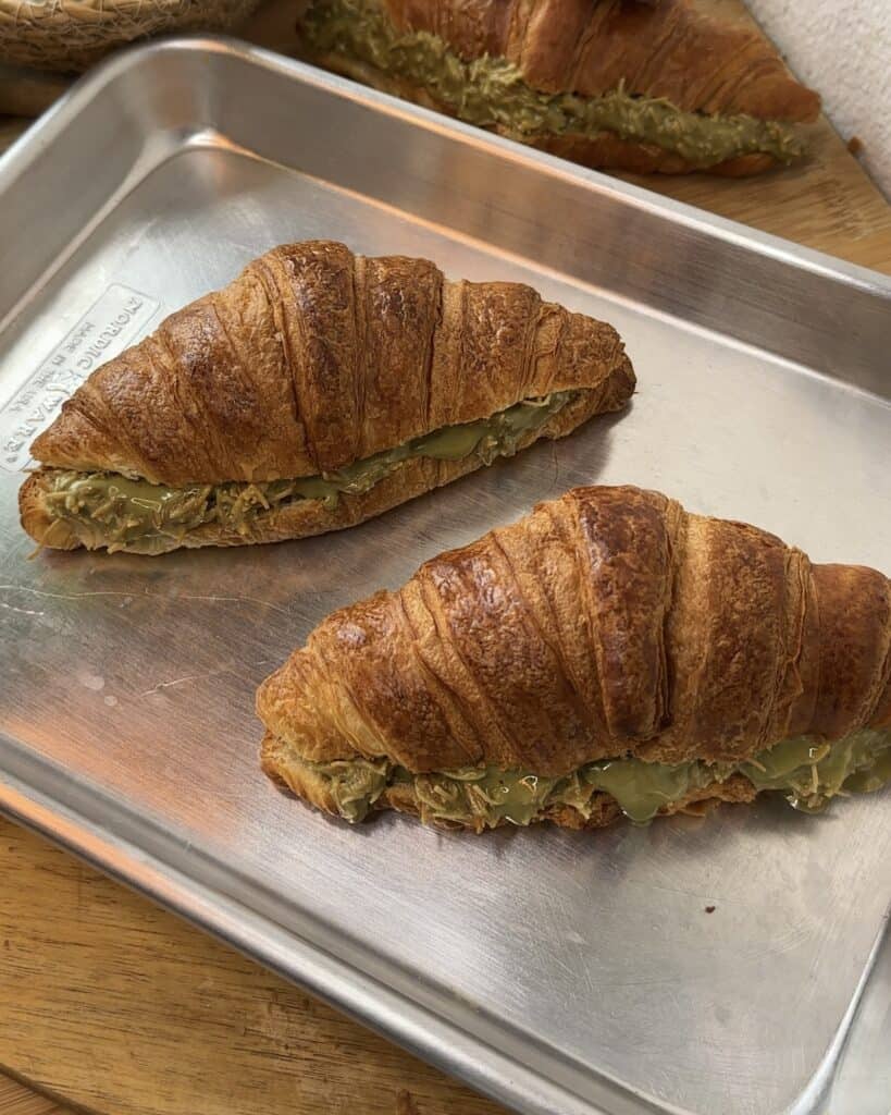 "Freshly baked croissants generously filled with creamy pistachio spread, placed on a silver baking tray, ready for the perfect indulgence."