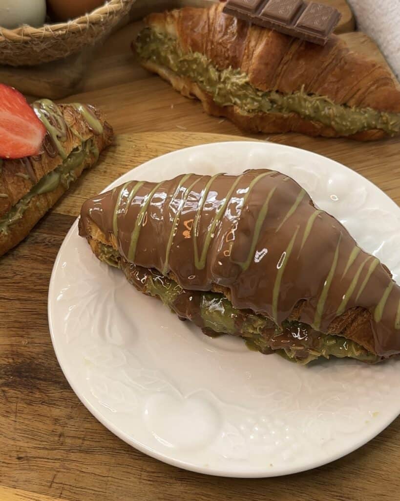 Decadent Dubai chocolate croissant filled with creamy pistachio spread, Kataifi with rich chocolate and a pistachio glaze, served on a rustic wooden board for an indulgent treat.