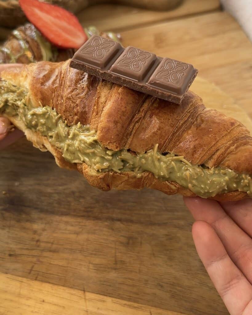 "Dubai chocolate croissant with a plain golden crust, filled with pistachio spread and toasted kataifi in the middle, topped with a piece of chocolate for an elegant finish."