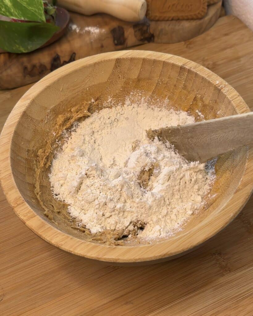 Image of a wooden bowl containing the mixed wet ingredients—softened butter, granulated sugar, brown sugar, and egg—along with the dry ingredients, including baking powder, flour, ground cinnamon, and ground ginger, being stirred together for the Biscoff marshmallow stuffed cookie dough.
