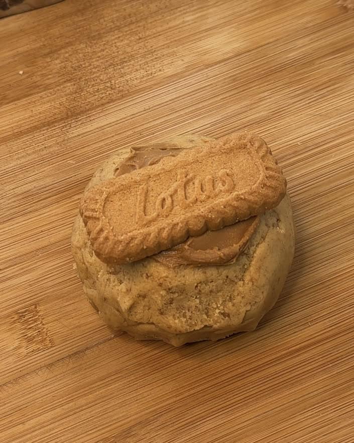 Image of a single-serve Biscoff stuffed cookie shaped into a round ball on a wooden board before baking and freezing, topped with a dollop of Biscoff spread and a Biscoff cookie for extra flavor.