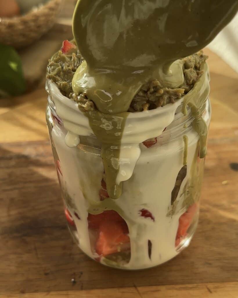 Close-up of the layers in a White Chocolate Dubai Strawberry dessert, with the top layer of toasted kataifi and pistachio cream. A spoon adds a dollop of pistachio spread on top, bringing extra flavor to this delightful treat.