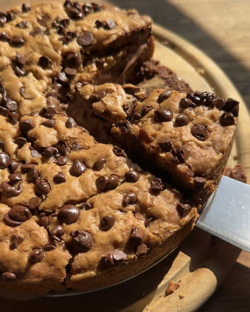 Close-up of a Nutella Stuffed Cookie Pie with a slice cut out, revealing the gooey Nutella center. The pie is golden brown with a soft, chewy texture, and the slice shows the layers of cookie dough and Nutella filling.
