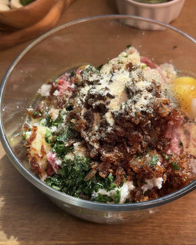 A glass bowl filled with ground beef, spices, caramelized onions, eggs, and fresh parsley, ready to be mixed for meatballs.