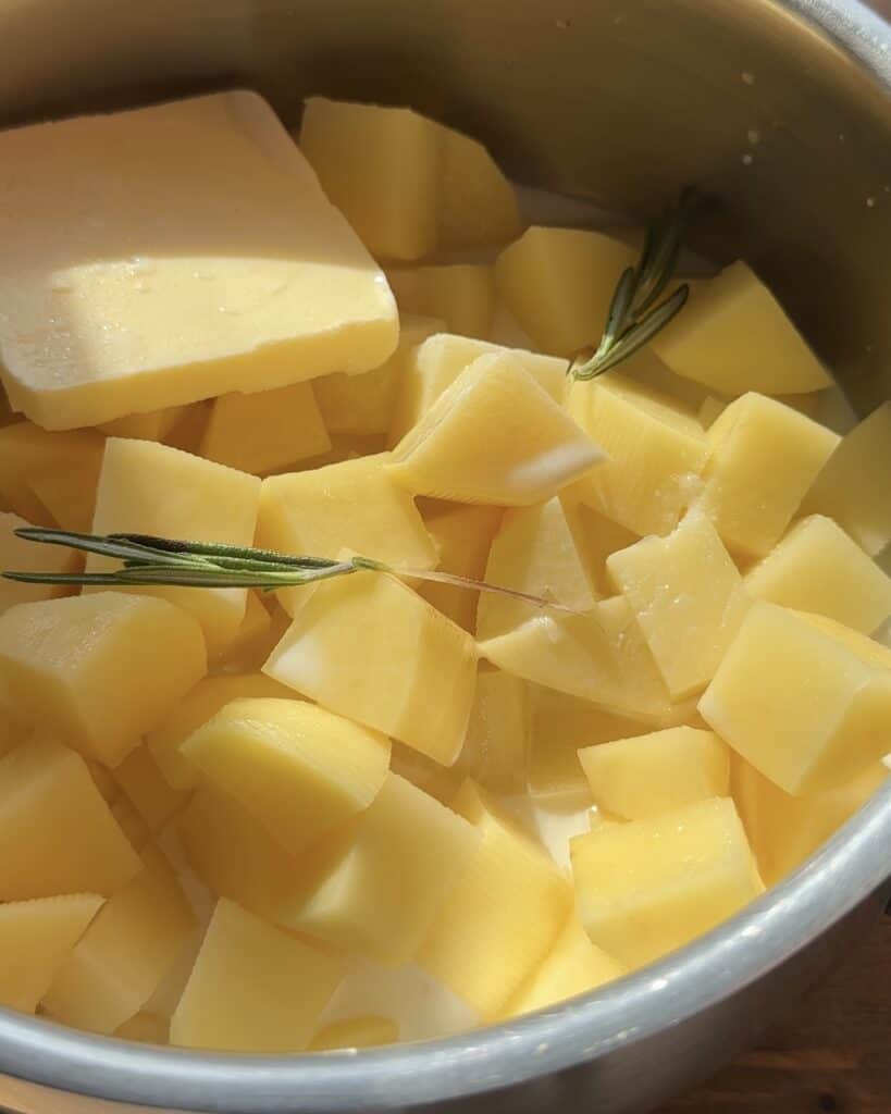 A pot filled with cut potatoes, fresh rosemary, and unmelted butter, prepared for boiling and cooking.